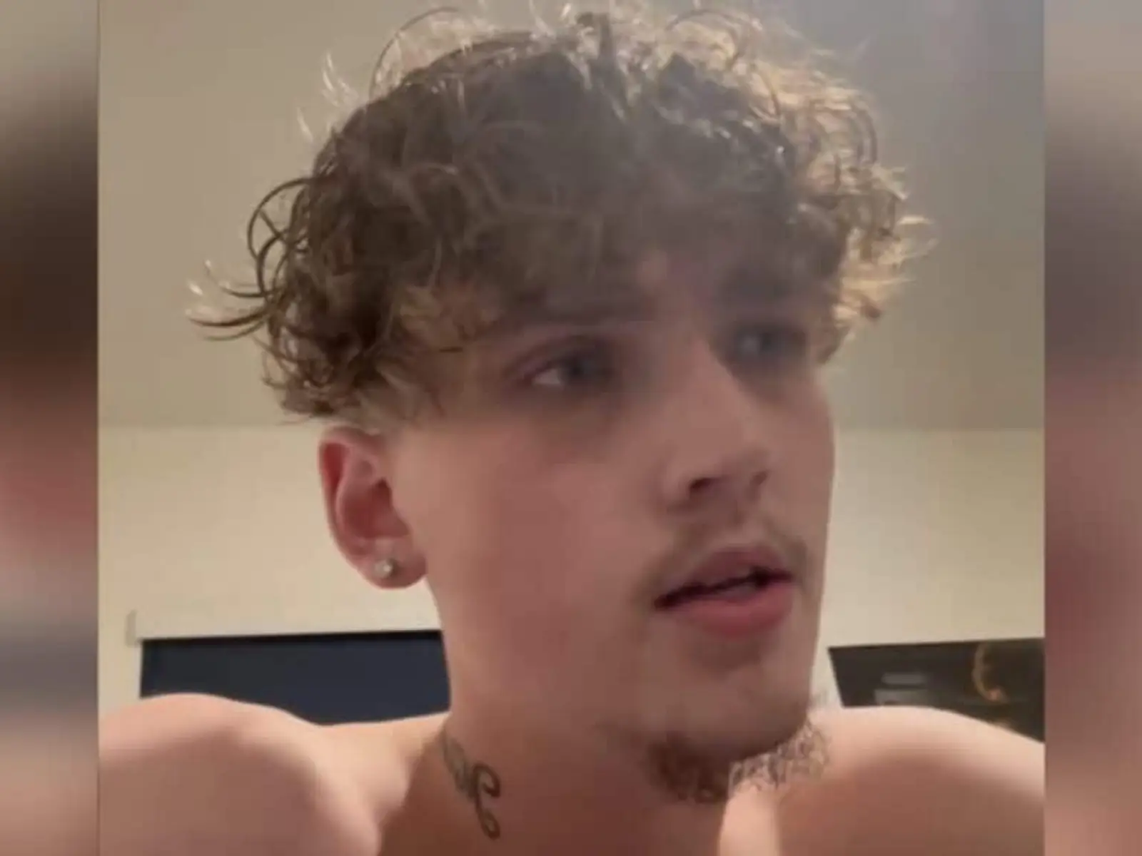 Close-up of a shirtless young man with curly hair and a stud earring, looking off to the side, with a small neck tattoo visible.