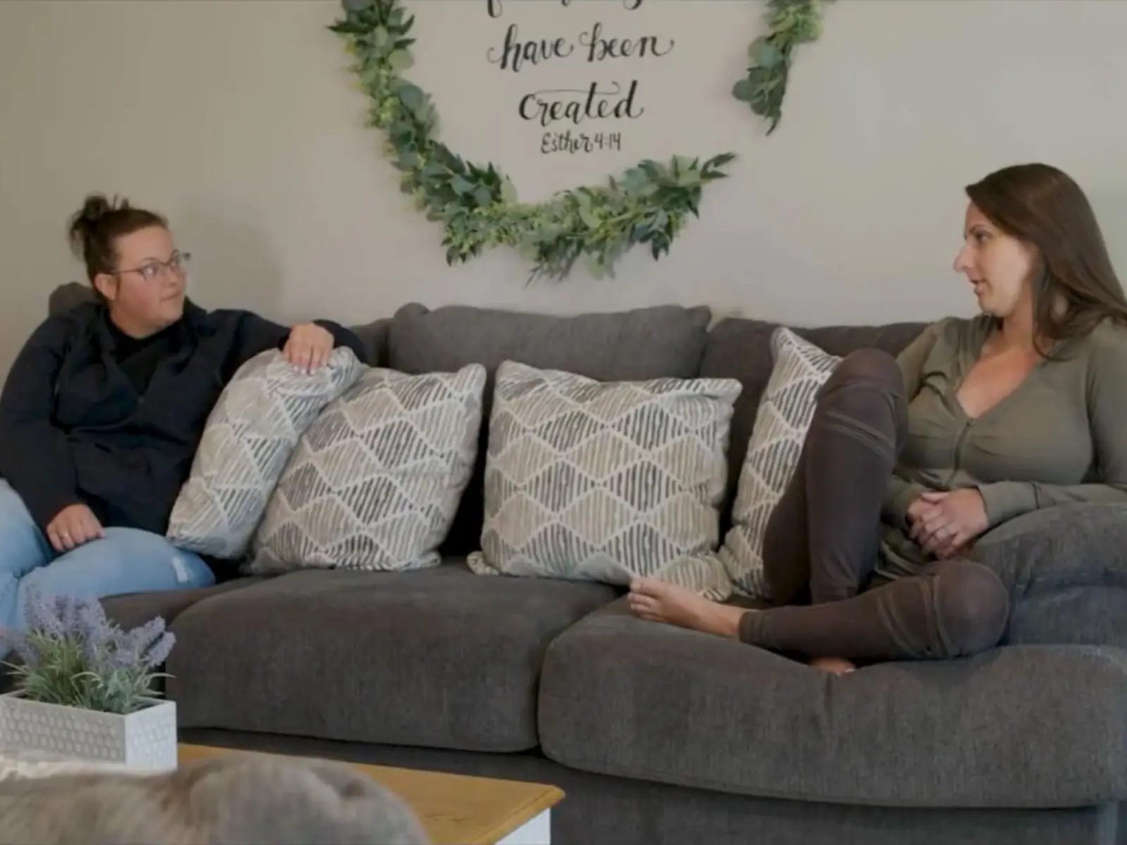 Two women sit on a gray sectional sofa facing each other, chatting in a cozy living room with a greenery wreath and wall quote above them.
